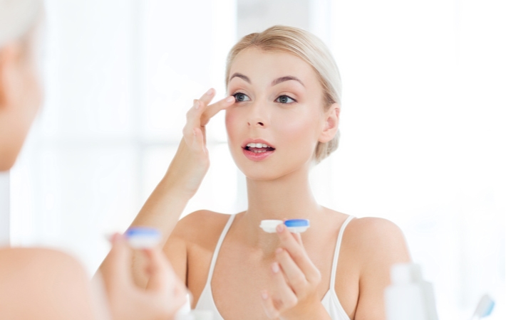 A woman putting in a contact lens in front of her bathroom mirror while holding the contact lens container in her other hand