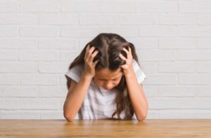A young girl hunched over on a wooden desk holding her head because she has head pain due to impaired vision