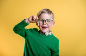 A little boy with a green shirt smiling while he is holding his green glasses on a yellow background