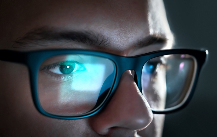 A close up of a man's glasses with the reflection of the screen he's looking at in the lenses