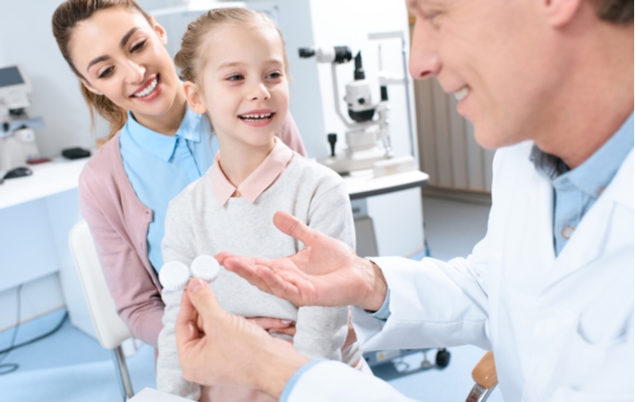 An optometrist holding a contact lens container, discussing them with a young girl and her mother