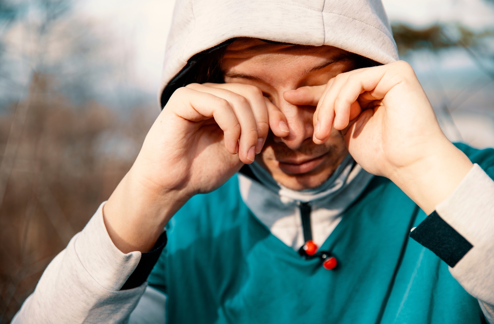A man outside wearing a hoodie with the hood up, rubbing his eye's because his eyes are sensitive to light