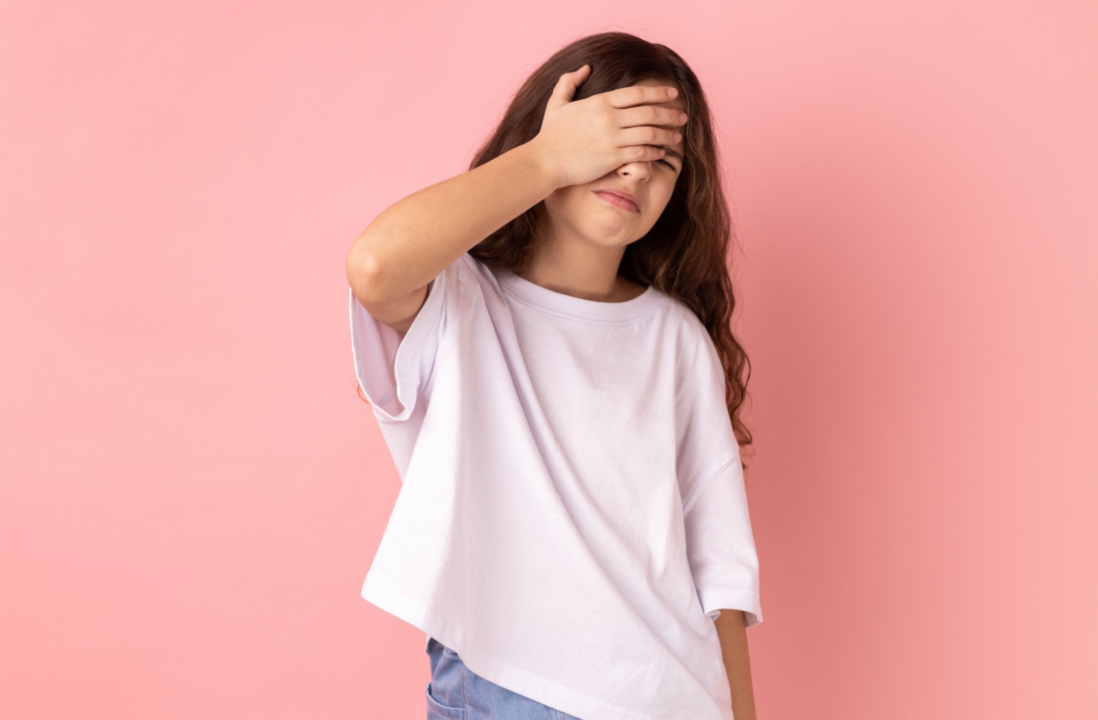 Against a pink background, a preteen in a white shirt covers one eye while grimacing in pain.