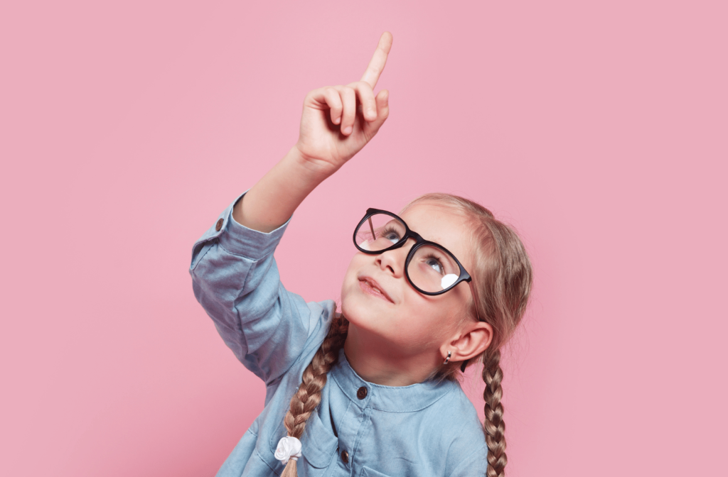 A young girl wearing glasses, sitting on the ground as she points up in the air above her