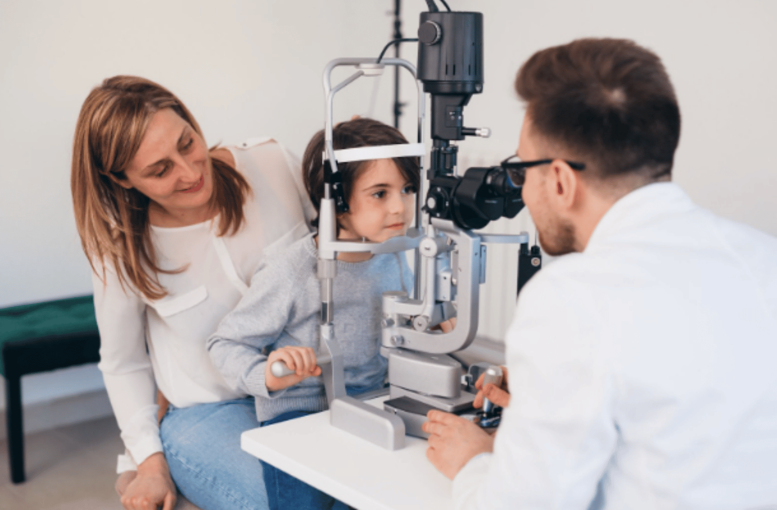 A male optometrist examines a little boy's eyes while the mother smiles.