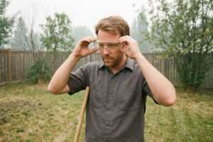 Adult in a grey work shirt putting on clear safety goggles in a fenced grassy yard, wooden tool handle at their side.