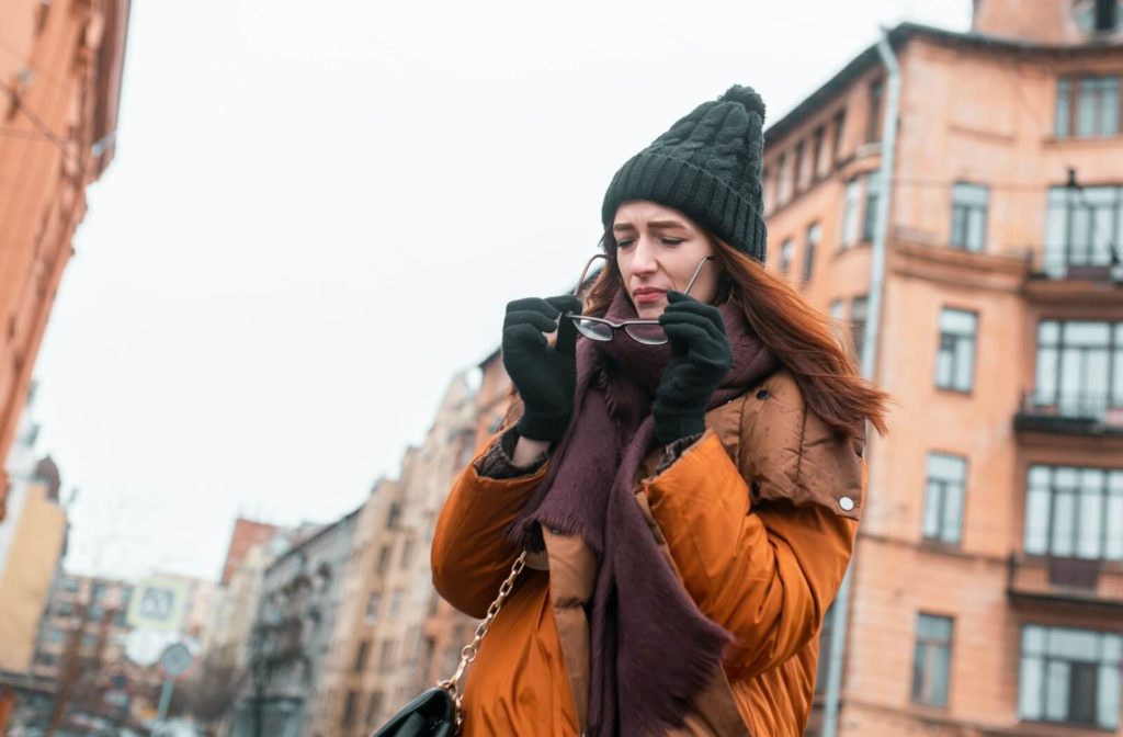 A young adult walking through the city in wintertime, taking off their glasses and squinting in frustration due to dry eyes.