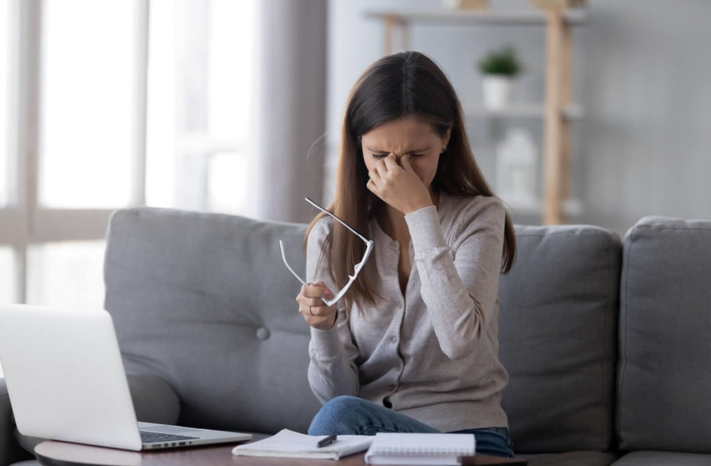 An adult working on a laptop, sitting on the couch, and taking off their glasses to rub their eyes due to wintertime dry eye discomfort.