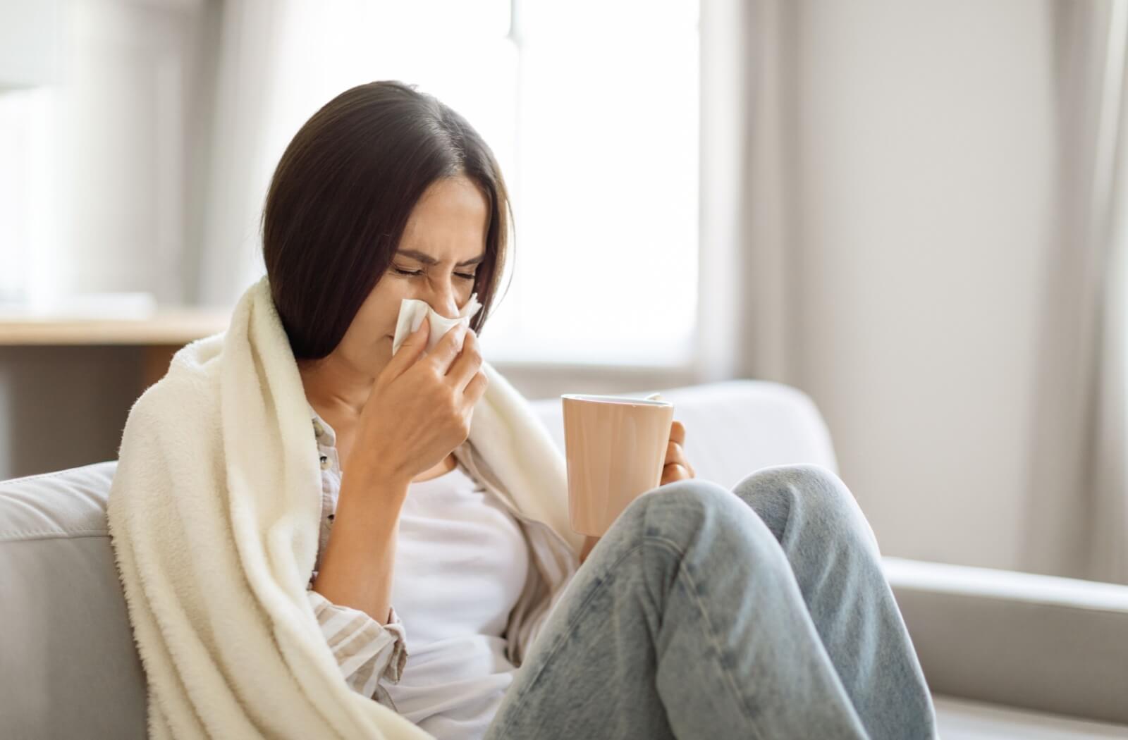 An adult drinking a tea blows their nose as they deal with a sinus infection.