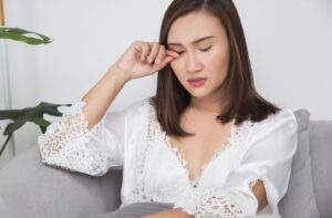 A person sitting on a couch closes their eyes and rubs one eye with their hand, showing signs of dry eye.