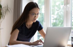 A person wearing glasses works on a laptop at a desk near a bright window.