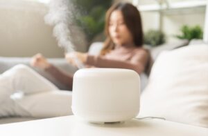 “White humidifier releasing mist on a table with a person relaxing on a couch in the blurred background.