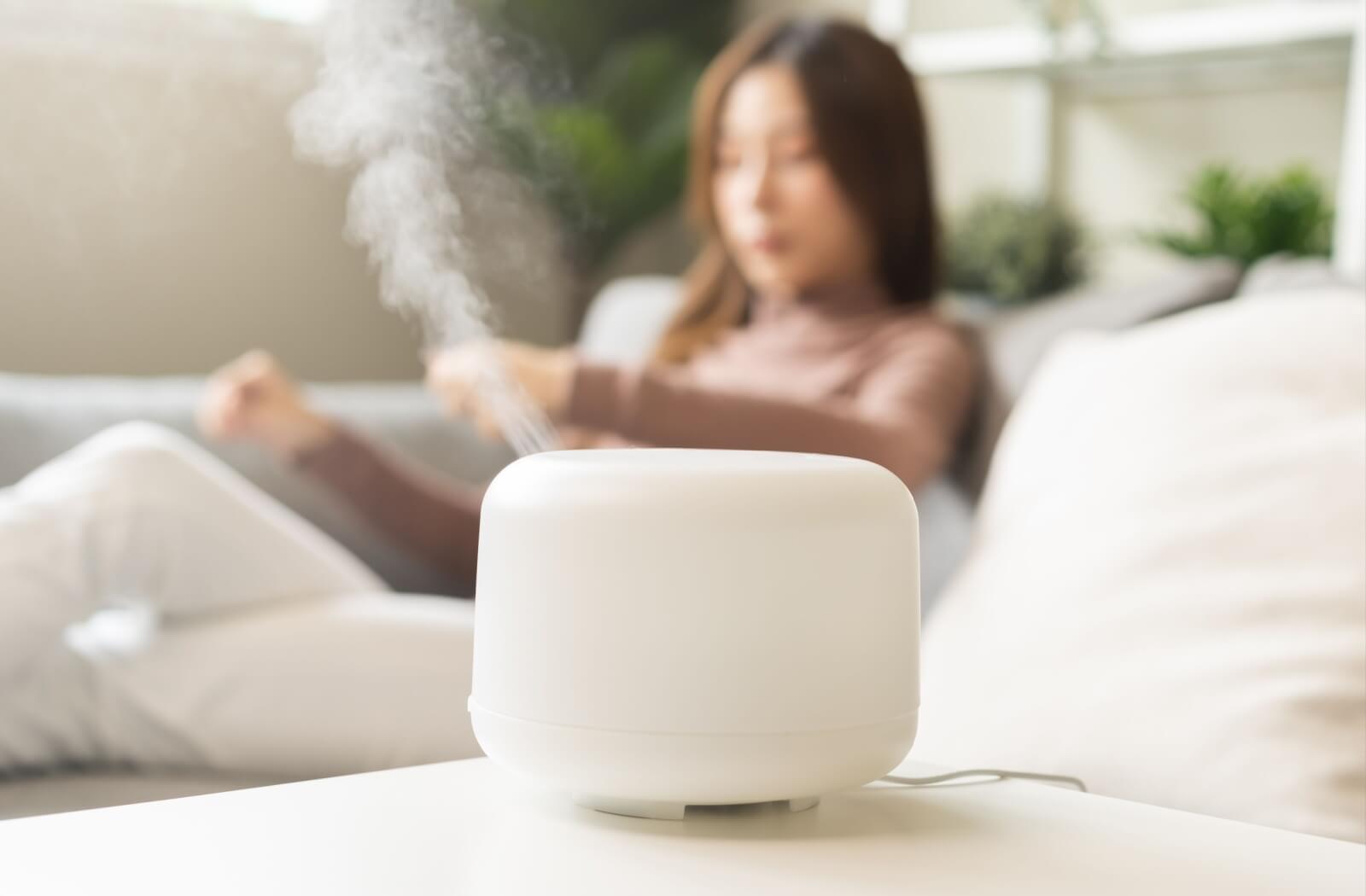 “White humidifier releasing mist on a table with a person relaxing on a couch in the blurred background.