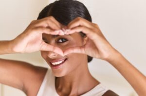 Smiling person forming a heart shape with their hands, symbolizing self-care and confidence.
