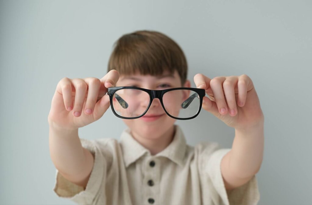 A child holding up a pair of black-framed eyeglasses toward the camera against a plain gray background.