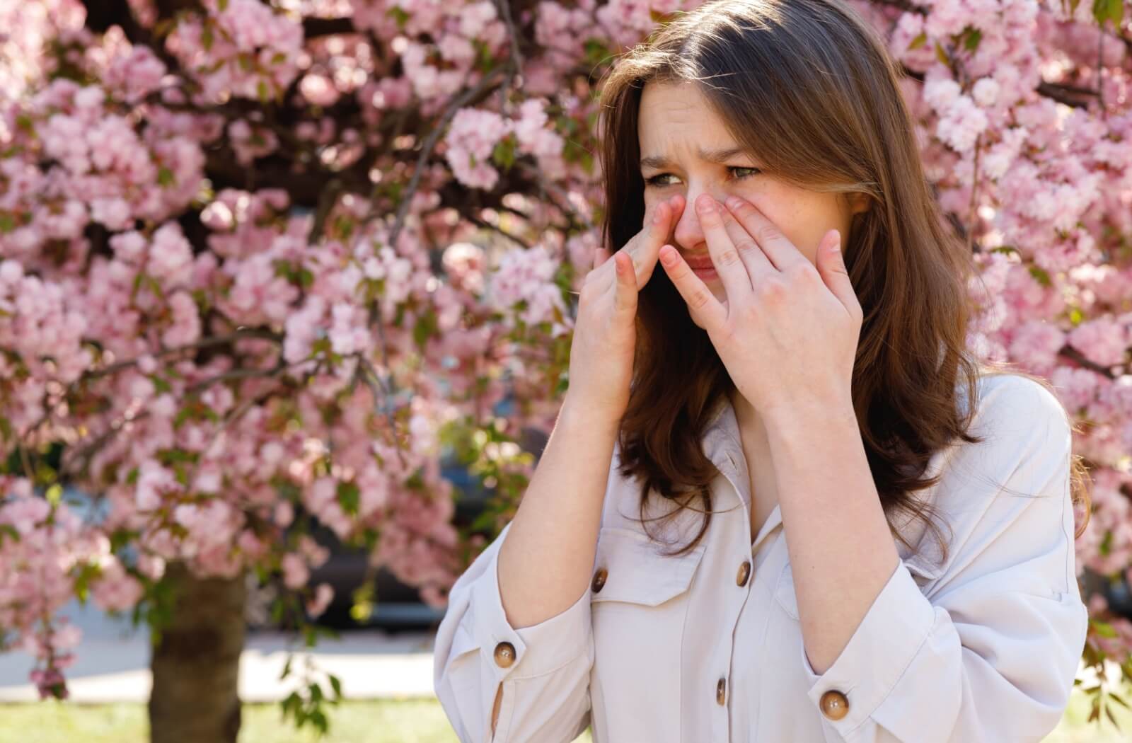 A young woman rubbing her itchy, swollen eyes while standing outdoors in front of a blooming cherry blossom tree.